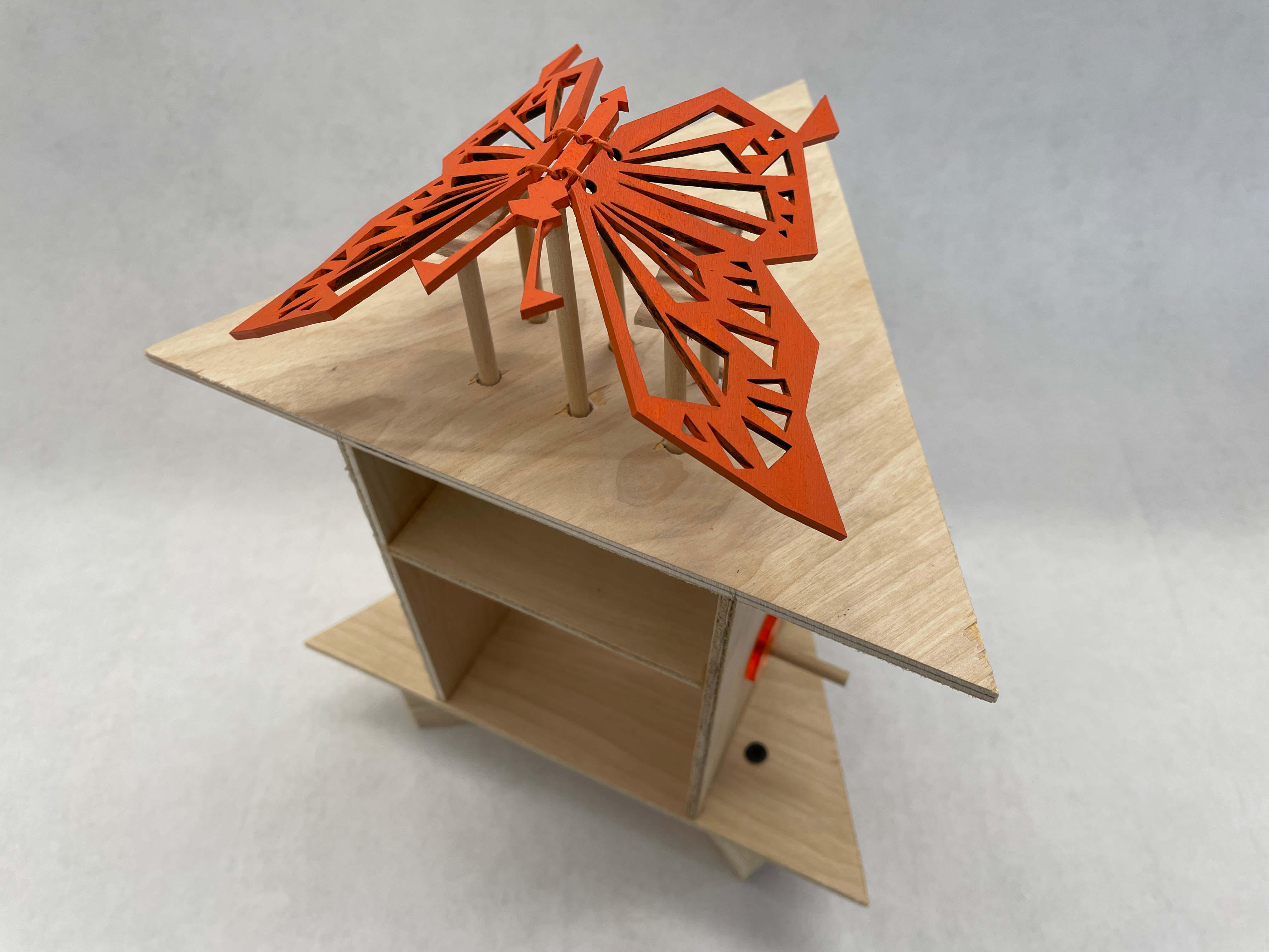 an image with a top slightly angled view of the orange butterfly automata which has an orange butterfly on top of a wooden triangular base with a dowel control in the middle